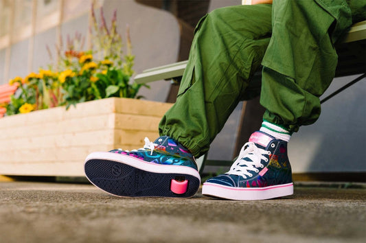 Alt text: Person wearing green pants and colourful Heelys roller shoes sits on a bench next to a flower planter, with one Heelys wheel visible under the sole of the right shoe.