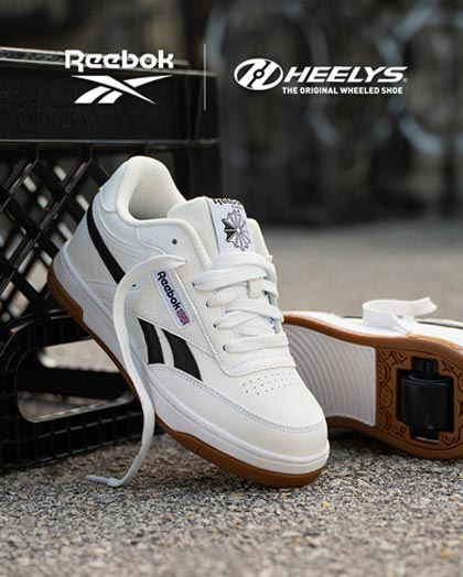 A pair of Reebok x Heelys roller shoes in white with black stripes and brown gum soles, displayed on a textured concrete surface with a black crate in the background. The Reebok and Heelys logos are visible at the top of the image.